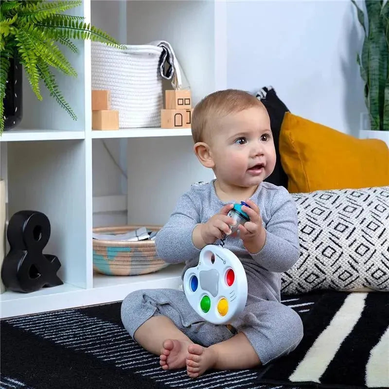 A baby in gray clothing sits on a black and white rug, holding a colorful Kids II Color Palette Popper sensory toy.