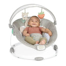 Baby sitting in the Ingenuity InLighten Baby Bouncer Seat, smiling, with toys hanging above and a soft gray seat.