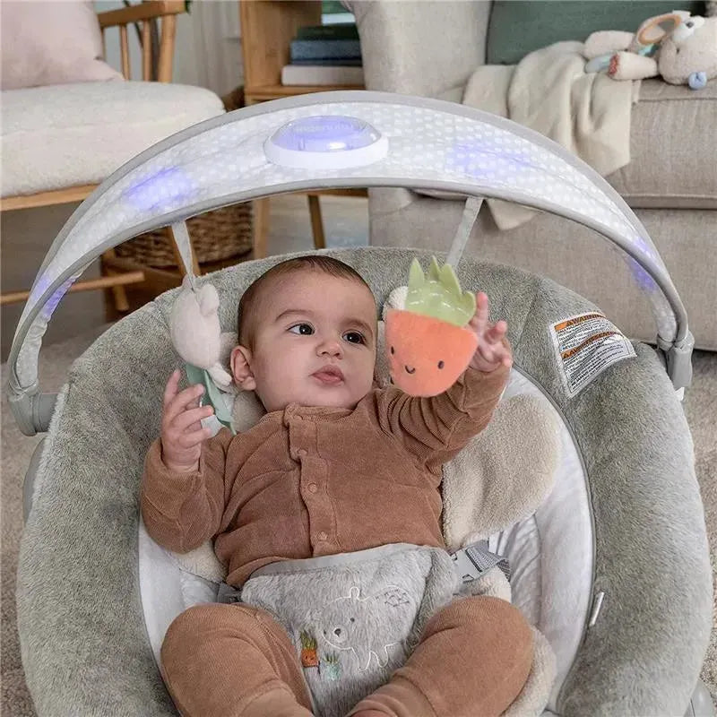 Baby in a brown outfit plays with a plush carrot toy in the Ingenuity InLighten Baby Bouncer Seat.