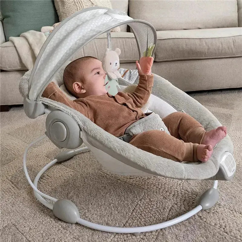 Baby seated in the Ingenuity InLighten Bouncer, interacting with hanging toys in a cozy living room setting.