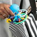 Blue octopus-shaped teether toy and rattle held in a hand, with colorful rings, against a striped bag.
