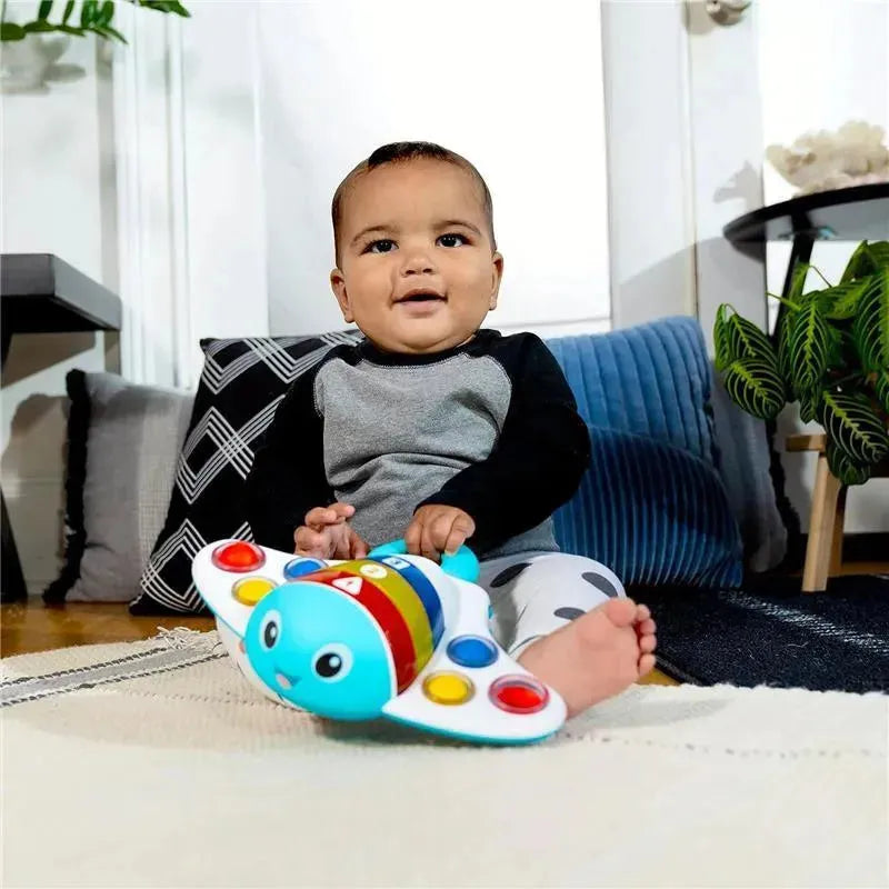 A baby sits on a rug playing with the Kids II Pop ‘N’ Explore Stingray Popper Toy, smiling. The toy has colorful buttons.