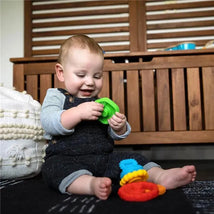 Infant sitting on a rug, smiling while playing with a green multi-textured teether toy, surrounded by colorful stacking rings.
