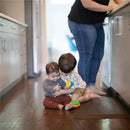 Two toddlers sit on a hardwood floor, with one child giving a gentle kiss to the other, who holds a colorful multi-textured teether toy. An adult stands in the background.