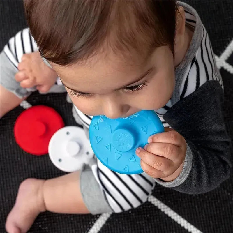 Infant interacting with a blue Kids II Stack & Wobble Zen Teether Toy, while red and white toys are visible nearby.