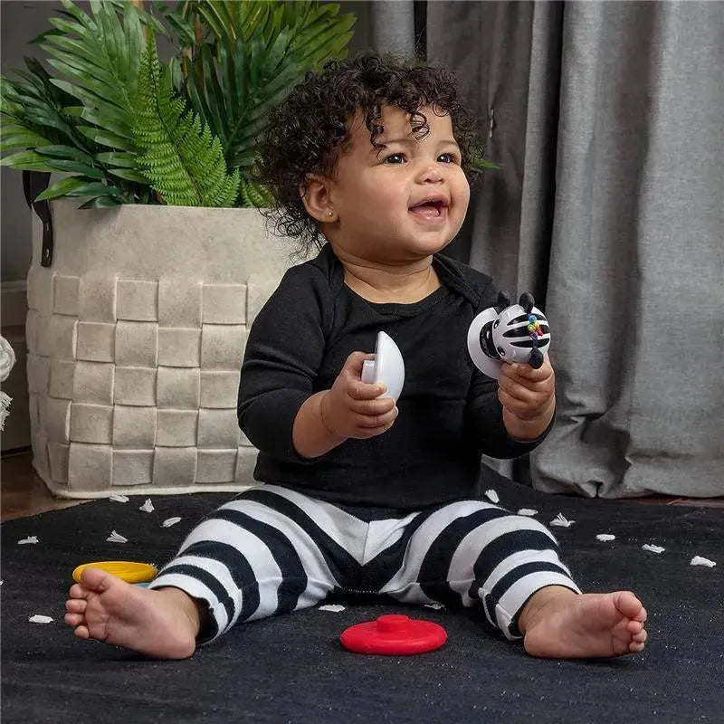 Toddler sitting on a black rug, holding a white teether and a zebra-patterned teether toy, smiling joyfully.