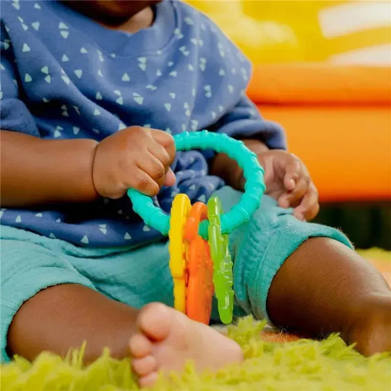 Colorful Kids II Tropical Chews teething ring held by a child, sitting on a green rug, with playful background colors.