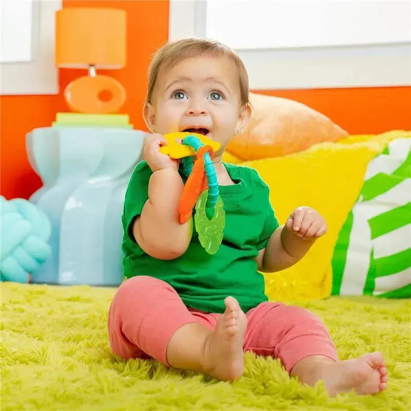 Toddler sitting on a green rug, happily biting a colorful Kids II Tropical Chews teething ring.