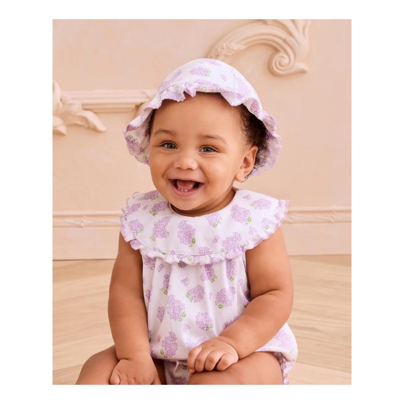 Smiling baby wearing a lilac floppy hat and a matching sleeveless outfit with floral patterns, sitting on a wooden floor.