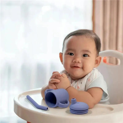 Kushies violet lilac Sili Sippy Cup with straw and lid, next to a smiling baby in a high chair.