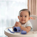 Kushies violet lilac Sili Sippy Cup with straw and lid, next to a smiling baby in a high chair.