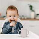 Baby with blue eyes sitting in a high chair, holding a gray spoon and next to a Kushies violet lilac sippy cup with a straw.