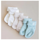 Three pairs of Kyte Baby socks in neutral colors: white, light gray, and sage green, each with a cuffed design.