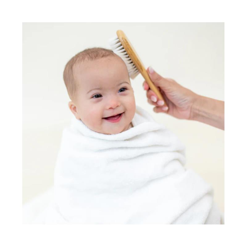 Smiling baby wrapped in a white towel, being brushed with a bamboo brush by an adult.