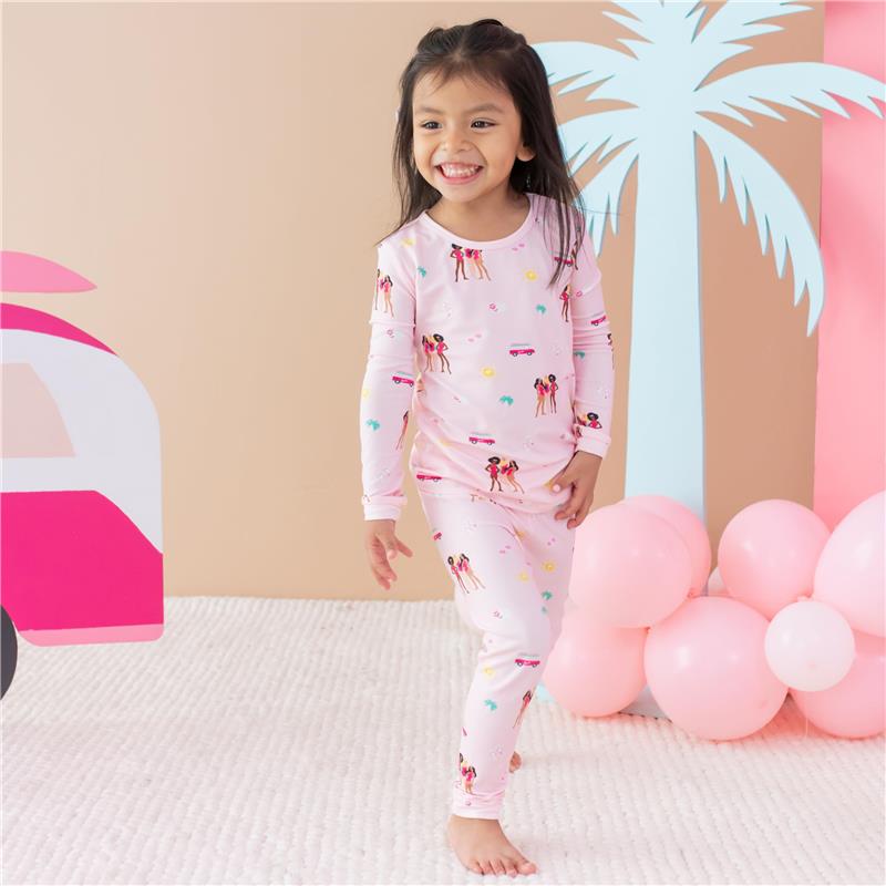 Baby girl wearing long sleeve pink pajamas with Barbie™ beach print, smiling while standing on a soft rug.