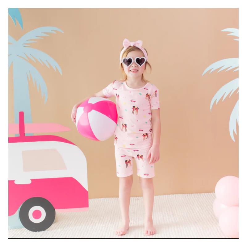 Baby girl in pink short sleeve pajamas with a Barbie™ beach design, holding a beach ball, smiling against a tropical backdrop.