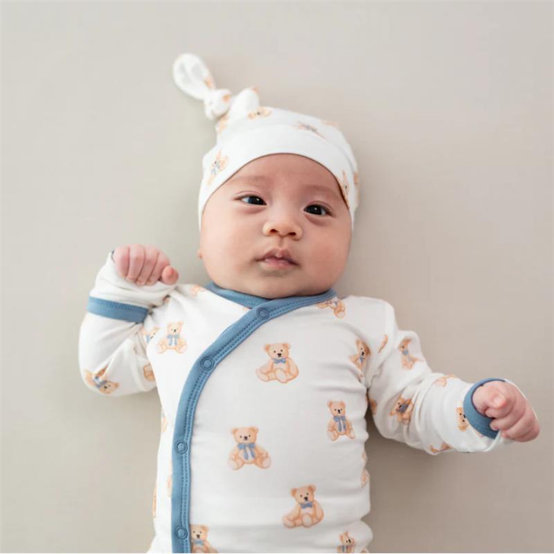 Baby girl wearing a teddy bear-print bodysuit with a matching cap, lying down on a light background.