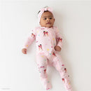 Baby girl in a pink zippered footie adorned with Barbie™ beach graphics and a matching headband, lying on a white surface.