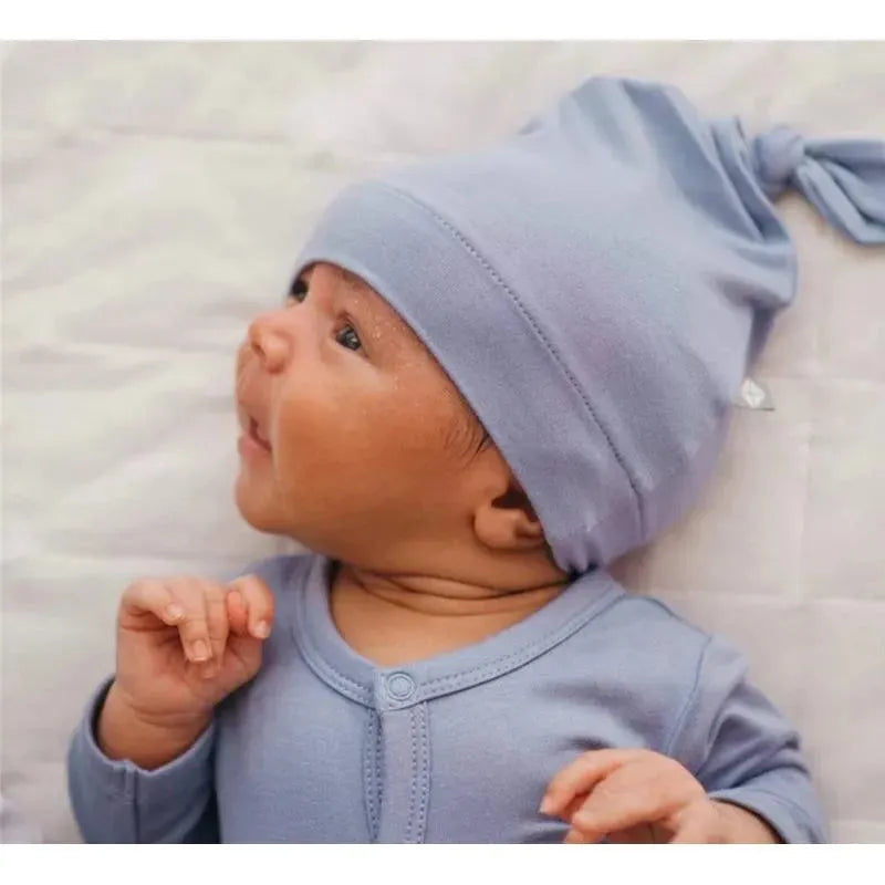 Bamboo baby knotted cap in slate worn by an infant lying on a soft surface, with the baby looking to the side.