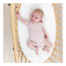 Baby wearing a soft blush Kyte Baby bodysuit, smiling while lying on a cozy white mattress in a woven bassinet.