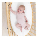Baby wearing a soft blush Kyte Baby bodysuit, smiling while lying on a cozy white mattress in a woven bassinet.