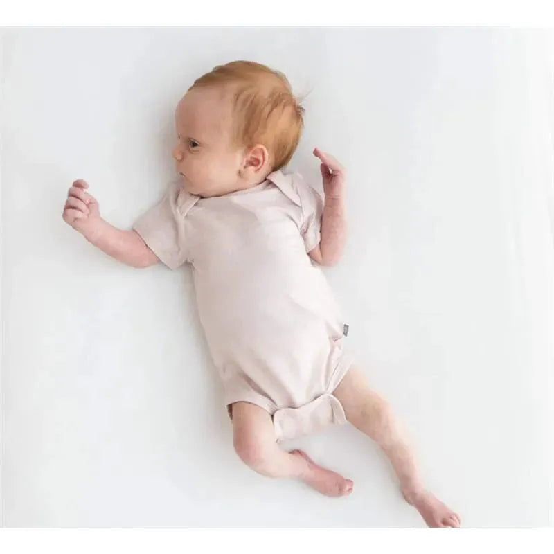Neutral blush baby bodysuit worn by an infant lying on a white surface, looking curiously to the side.