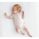 Neutral blush baby bodysuit worn by an infant lying on a white surface, looking curiously to the side.