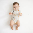 Baby wearing a Kyte Baby neutral bodysuit with a fluffy dog pattern, smiling and sitting on a white background.
