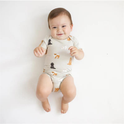 Baby wearing a Kyte Baby neutral bodysuit with a fluffy dog pattern, smiling and sitting on a white background.