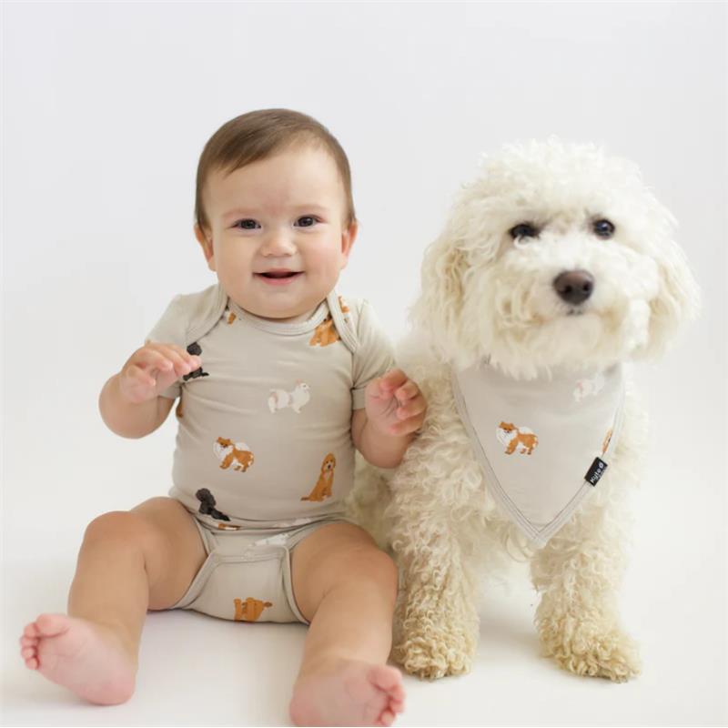 Baby wearing a neutral bodysuit with fluffy dog prints, sitting beside a fluffy white dog wearing a bandana.