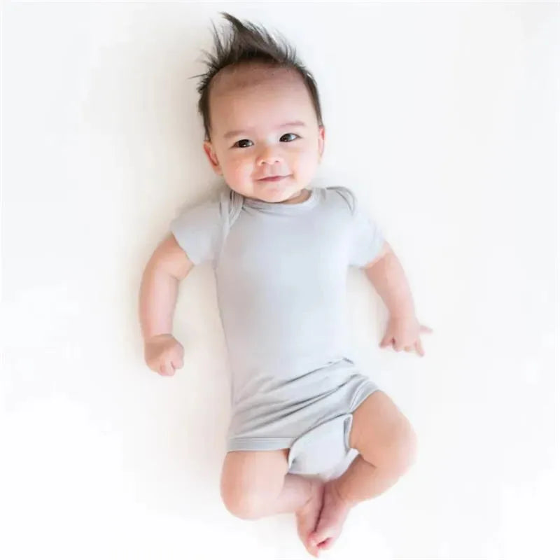 Neutral baby bodysuit in storm color worn by a smiling baby lying on a white surface.