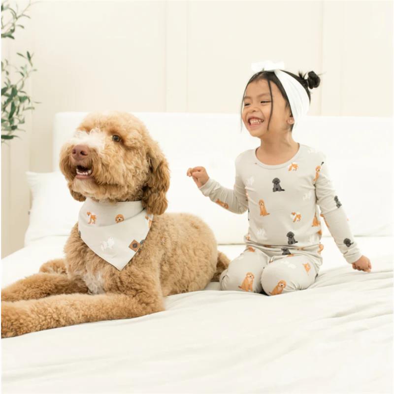A girl in long sleeve pajamas featuring fluffy dog patterns sits next to a dog wearing a matching bandana, both smiling on a bed.