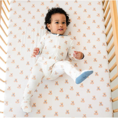 Baby wearing a Kyte Baby zippered footie with teddy bear print, lying on a crib sheet featuring teddy bear patterns.