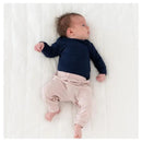 A baby lies on a white blanket, wearing a navy long-sleeve shirt and blush-colored pants with an elastic waistband.
