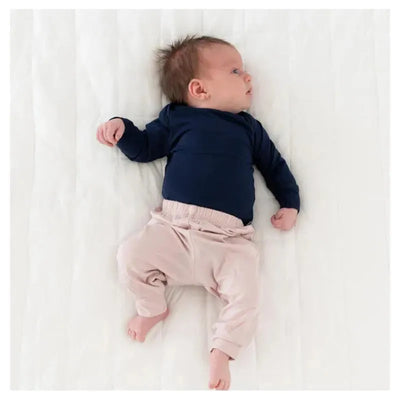 A baby lies on a white blanket, wearing a navy long-sleeve shirt and blush-colored pants with an elastic waistband.