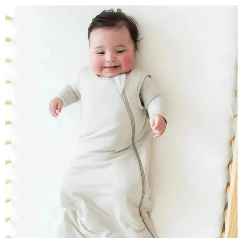 Bamboo sleep bag in oat color on smiling baby, laying on a soft surface, showcasing a zipper design and cozy fit.