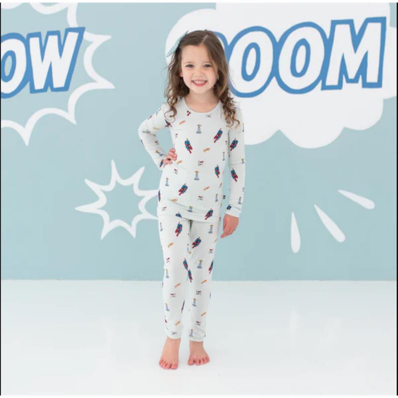 Toddler girl wearing Kyte Baby bamboo long sleeve Superman pajamas, smiling against a light blue comic-style background.