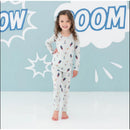 Toddler girl wearing Kyte Baby bamboo long sleeve Superman pajamas, smiling against a light blue comic-style background.