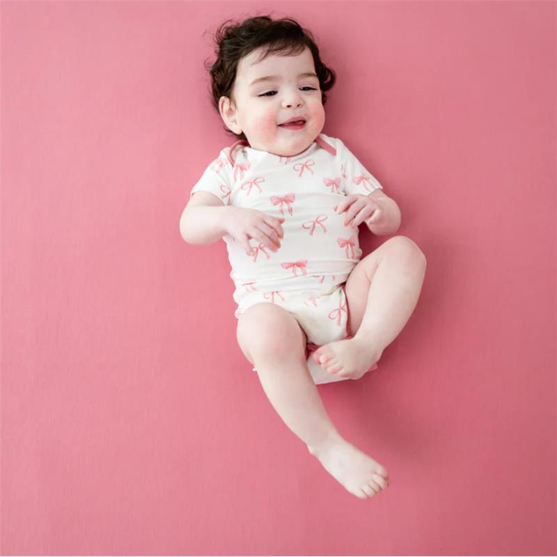 Infant wearing a white bodysuit with pink bow prints, smiling on a pink background.