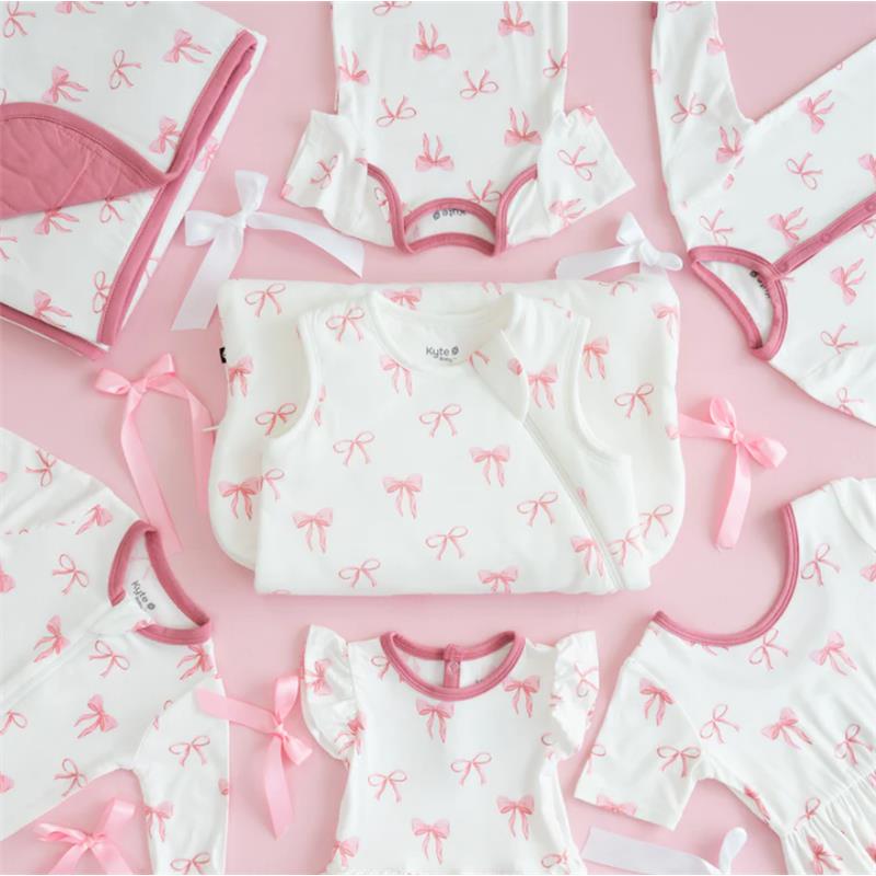 Kyte Baby bodysuits and a sleep sack, featuring a pink bow print on a white background, arranged on a pink surface.