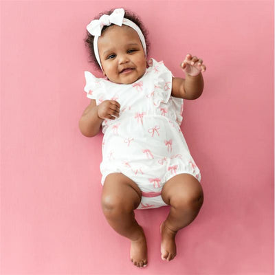 Kyte Baby Bubble Romper In Bow worn by a smiling infant, features a white fabric with pink bows and ruffled sleeves.