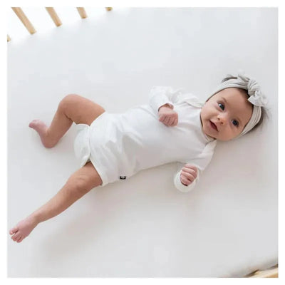 Baby lying on a crib mattress wearing a white long sleeve bodysuit and a matching headband, smiling at the camera.