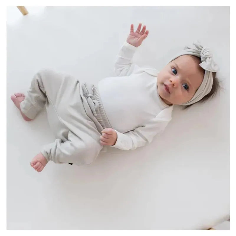 Long sleeve white bodysuit paired with light gray pants, worn by a baby with a matching bow headband, lying on a soft surface.