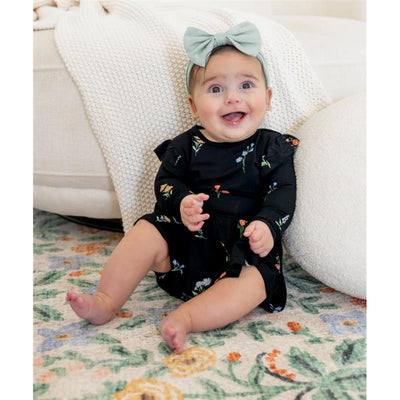 Baby wearing a black floral Kyte Baby long sleeve twirl bodysuit dress, smiling while sitting on a colorful rug, with a green headband.