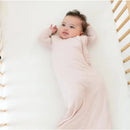 Bamboo sleep bag in blush color worn by a baby laying on a white crib mattress, with wooden crib sides visible.
