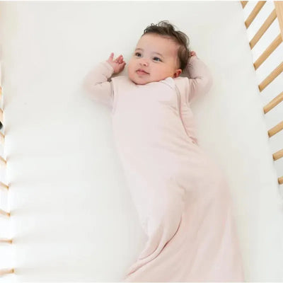 Bamboo sleep bag in blush color worn by a baby laying on a white crib mattress, with wooden crib sides visible.