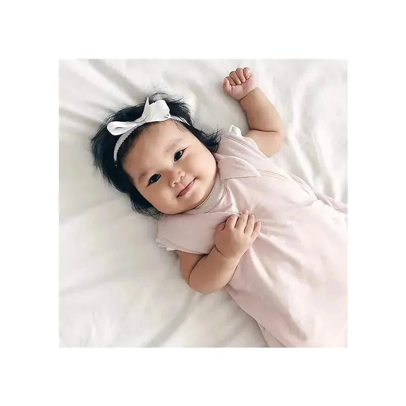 Baby smiling while lying on a bed, wearing a blush-colored outfit and a white bow headband.