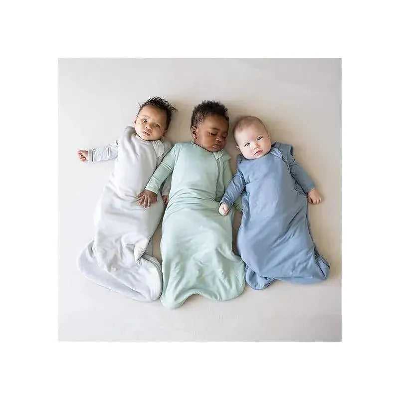 Three babies in colored Kyte Baby bamboo sleep bags: gray, green, and blue, lying on a light-colored surface.