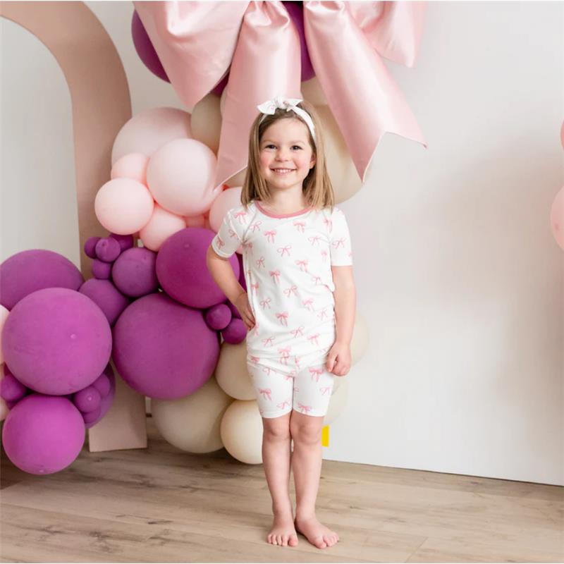 Short-sleeve white pajamas with pink bow prints, worn by a smiling girl standing in front of colorful balloons.