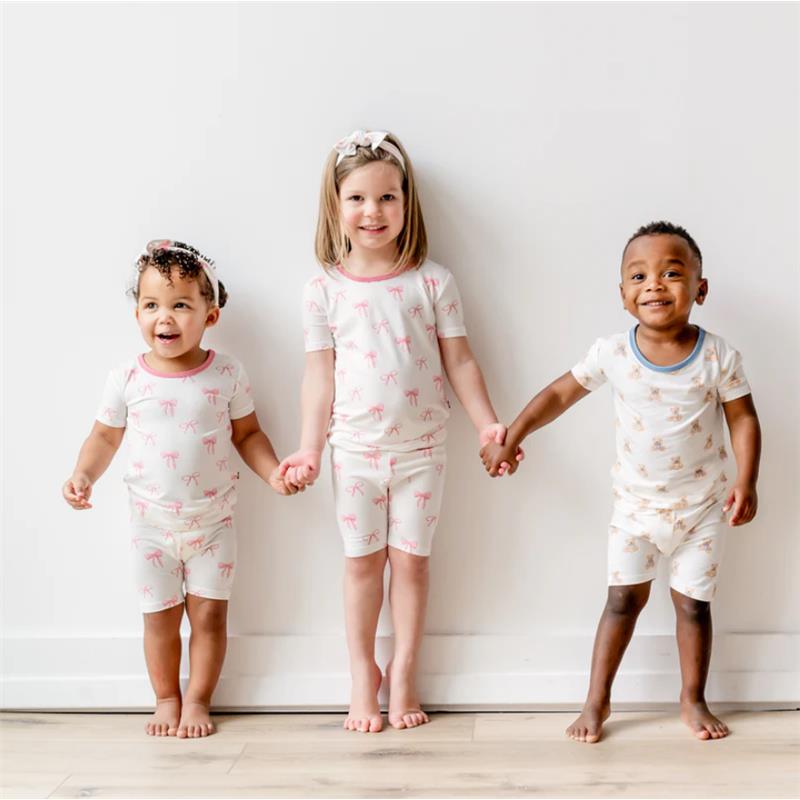 Three children wearing Kyte Baby short sleeve pajamas stand together, smiling. The pajamas feature playful prints and soft colors.